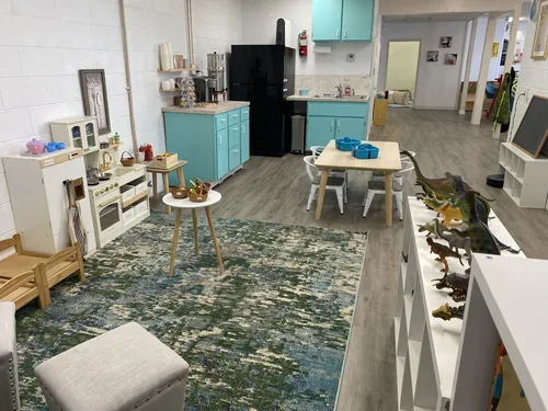 Children's play area with toy kitchen, small tables with baskets of toys, a rug, and toy dinosaur figurines on a white shelf in a classroom setting.