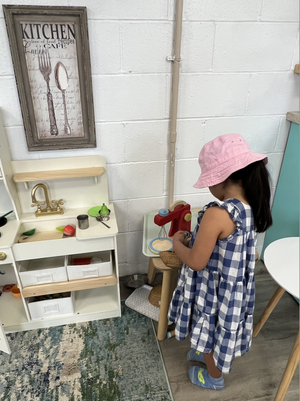 A young girl wearing a pink hat and a blue checkered dress playing with a toy red scale in a play kitchen area.