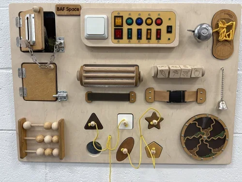 Interactive sensory board with various wooden and metal objects including knobs, switches, a bell, a star-shaped block, an abacus, and other tactile elements for children.