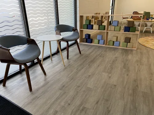 Interior of a modern space with two gray chairs, a small white round table, a wooden partition with upholstered cubes, and tables with art supplies in the background.