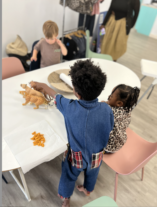 Children playing with animal figurines around a white table with a paper towel and orange toy pieces in a classroom.