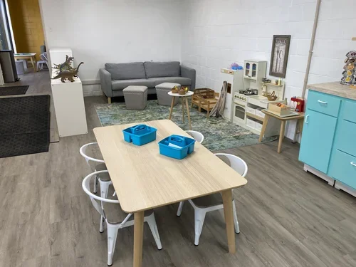 A children's play area with a wooden table and six white chairs, two blue plastic bins on the table, a gray sofa with an ottoman, a small rug, a play kitchen set, and dinosaur toys on a white pedestal.