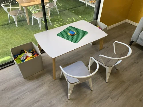 A white table with a green Lego baseplate and a small Lego structure on top, two chairs with white frames and wooden seats around the table, and a box of assorted Lego pieces on the floor.