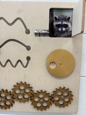 A wooden activity board with gears and a small window showing a photo of a raccoon.