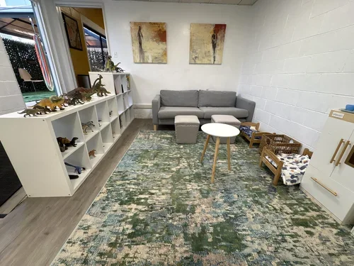 Living room with white brick wall, gray sofa, colorful abstract paintings, toy dinosaurs on a white shelf, and a multicolored area rug.