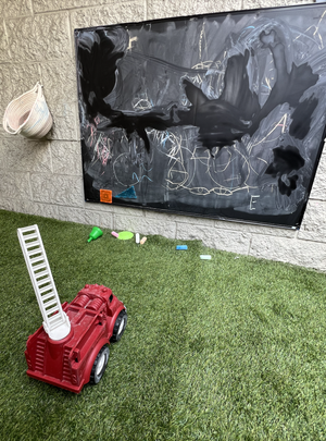 Outdoor play area with green grass, a red toy fire truck, scattered chalk pieces, a white ladder, a green toy, and a large black chalkboard mounted on a brick wall, featuring abstract black and white drawings.