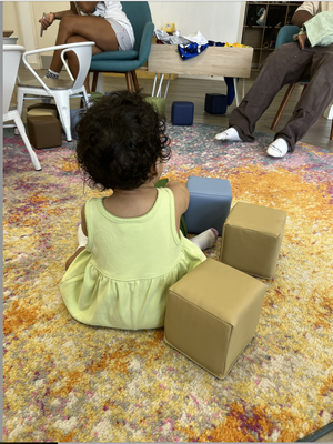 A young girl with curly dark hair wearing a yellow dress sitting on a colorful patterned carpet, playing with blue and beige foam blocks.