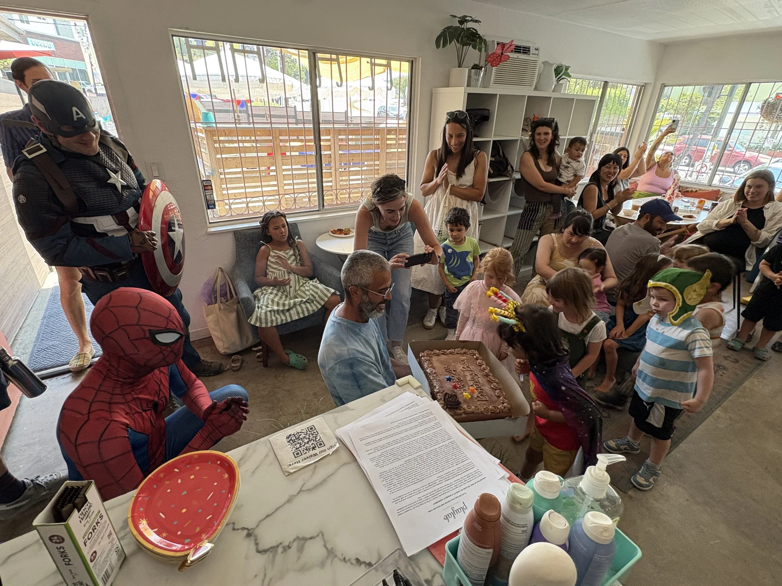 Children and adults gathered in a bright room celebrating a children's birthday with a cake, balloons, and party decorations. Some people are dressed in superhero costumes, including Spiderman and Captain America. The room has large windows, and there are tables with party supplies and a cake on a table at the front.