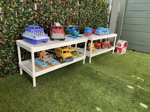 Outdoor children's play area with artificial grass, a small table with chairs, hanging fabric curtains, and toy vehicles on white shelves, enclosed by a privacy fence and a roof.