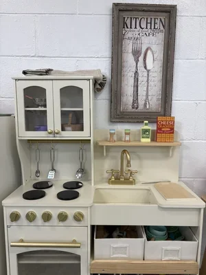 A children's play kitchen set with a white and beige color scheme, featuring a stovetop, oven, shelves, small containers, and utensils, against a white brick wall with a framed wall art piece.