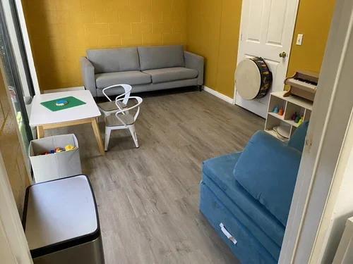 Playroom with gray and blue couches, a small white table with a green Lego baseplate, plastic building bricks, a white bookshelf with colorful toys, a large drum, and a small wooden piano against mustard yellow and white walls.