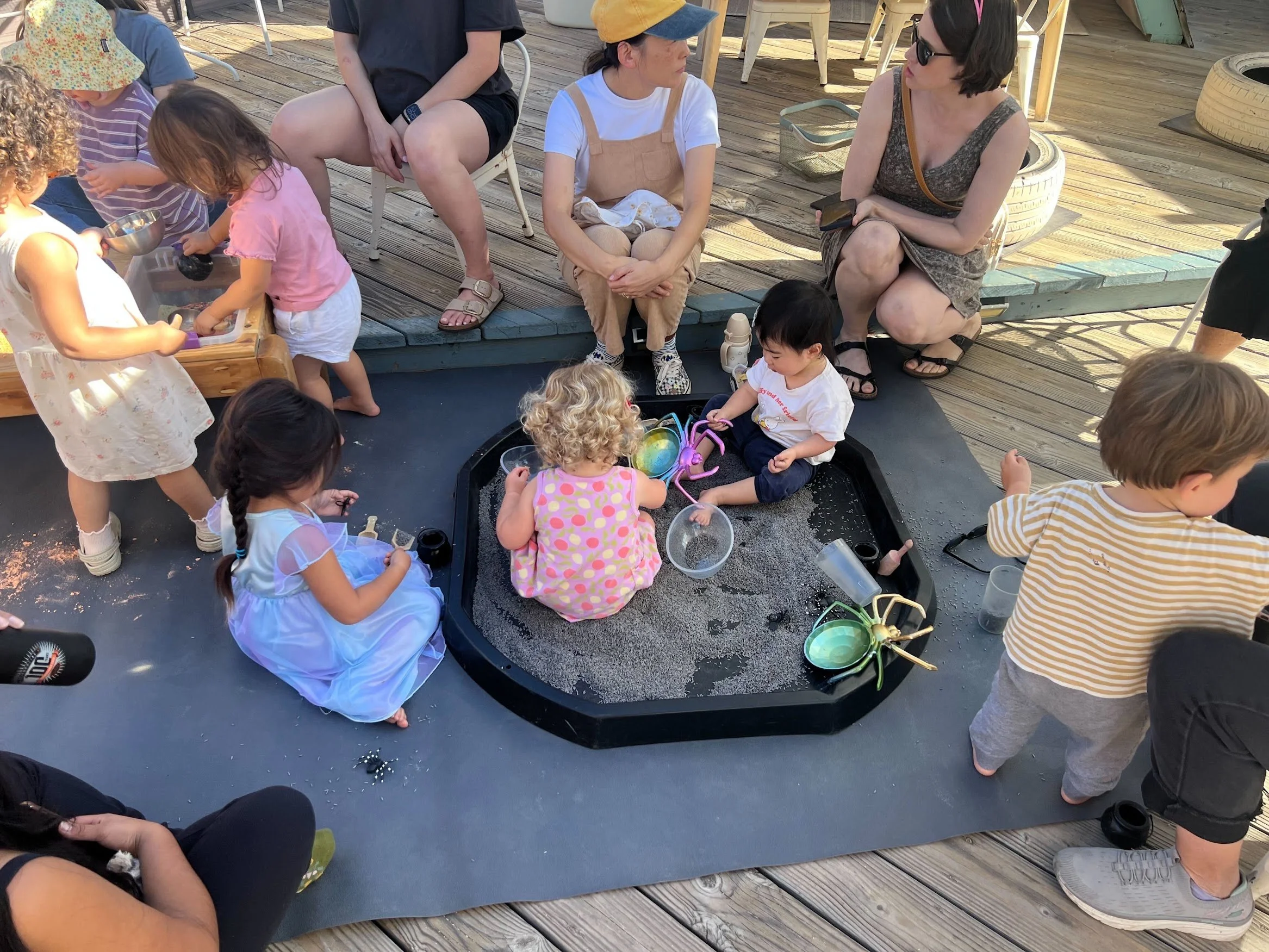 Children exploring a sensory play tray with sand and toy spiders on the outdoor deck at PlayLab