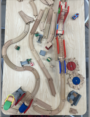A wooden train track set assembled on a light-colored table, including curved and straight tracks, train cars, and accessories like bridges and signal flags.