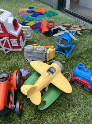 Playroom toys scattered on green artificial grass, including a red and white barn, toy vehicles, plastic blocks, and a toy dog.