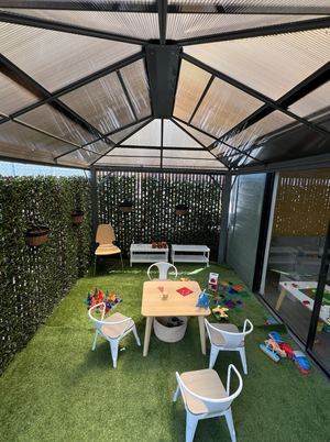 Children's outdoor play area with a small table and chairs, toy blocks, and a shaded, enclosed space with artificial grass and decorative plants.