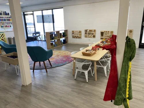 Children's play area with colorful chairs, a round rug, toy shelves, wall-mounted activity boards, a table with toy trucks, and two plush dinosaur costumes hanging on a pole.