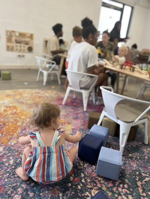 A young child with short brown hair and a colorful striped dress sits on a multicolored rug, playing with foam blocks in a room filled with adults and children sitting at a table.