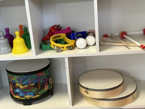 Children's musical instruments stored on a white shelf, including colorful bells, maracas, mallets, and tambourines.