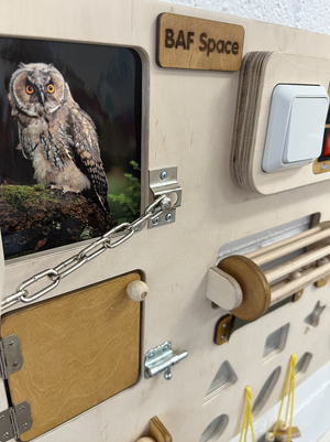 Close-up of a wooden activity board with a printed picture of an owl, a small wooden cabinet, a chain, a door latch, and various wooden and metal cladding features. A small wooden sign reads 'BAF Space'.