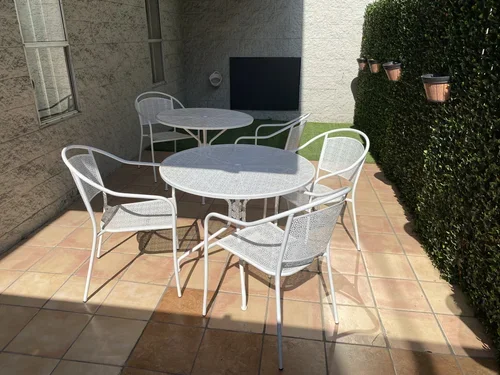 Outdoor patio with a round metal table, six matching metal chairs, a brick wall, green hedges with small flower pots, and a television mounted on the wall.