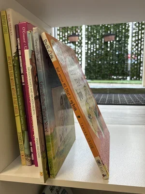 A collection of children's books displayed on a white shelf with titles visible, including 'Karen' and 'Charlie and Lola', along with a tea party-themed book titled 'Tea & Cake'.