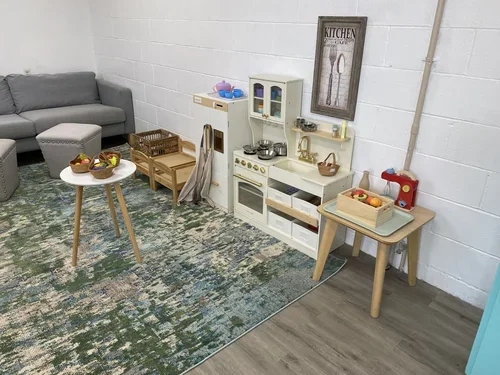 A children's play kitchen set with a white and beige color scheme, featuring a stovetop, oven, shelves, small containers, and utensils, against a white brick wall with a framed wall art piece.