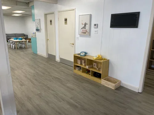 Interior of a children's play area with a hallway, two closed bathroom doors, a small wooden shelf with toy phones on top, and a play area with a table and chairs in the background.