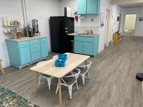 A small cafeteria or break room with a wooden table and four white chairs. The table has blue plastic containers on it. To the left, a turquoise cabinet with coffee and tea supplies.