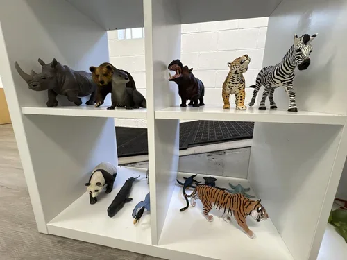 Collection of various animal figurines on a white shelving unit including a rhinoceros, bear, otter, tiger, zebra, panda, and other animals.