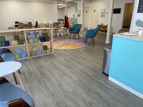 Interior of a colorful daycare or playroom with small tables, chairs, a bookshelf with blocks, a round colorful rug, and cozy chairs.