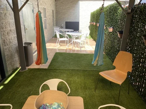Outdoor patio area with a wooden table, four white chairs, a large woven basket on the table, and an artificial grass surface. Behind a glass sliding door, an indoor dining table with white chairs and a toy on the table are visible.