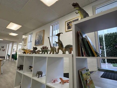 Shelf with various toy dinosaurs and animals inside a room with framed artwork on the wall and books on a nearby bookshelf.