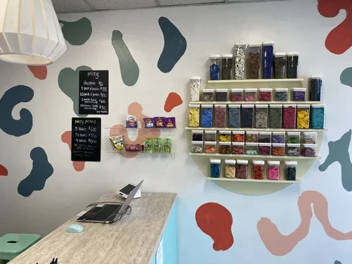 Cake decorating shop wall with colorful sprinkles containers, pricing signs, and a counter with a laptop.