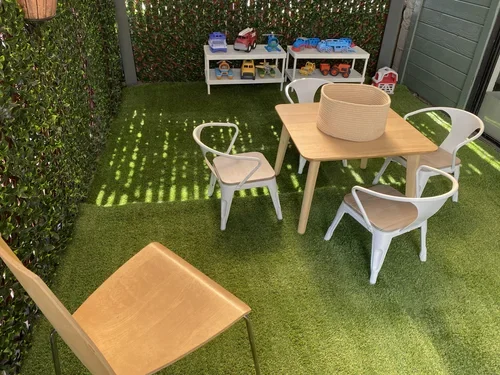 Outdoor play area with a wooden table, four white chairs with wooden seats, a small shelf with toy vehicles, and a wooden chair, on artificial grass with a hedge and a small shed in the background.