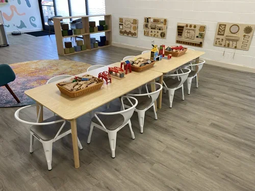 A playroom with a long wooden table holding baskets of wooden train toys, surrounded by white chairs, and decorated with educational wall panels. There is a colorful rug and a shelf with colored blocks, with a door and window in the background.