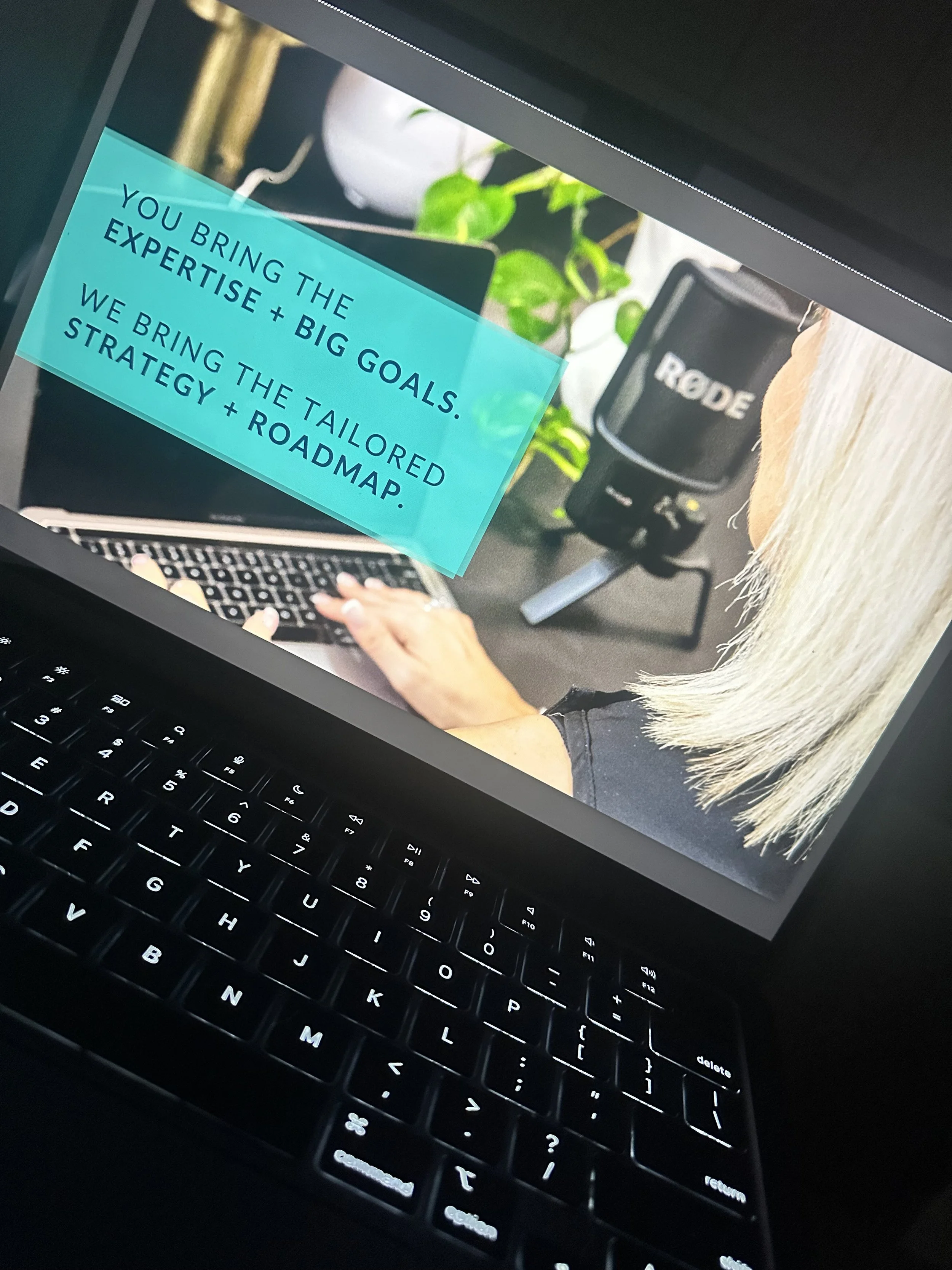 A computer screen displaying a presentation slide with text that says, 'You bring the expertise + big goals. We bring the tailored strategy + roadmap.' The slide also shows part of a laptop keyboard and a person with blonde hair working on the laptop, a Rode microphone, and a green leafy plant in the background.
