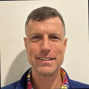 A man with short brown hair and a slight beard smiling at the camera against a light-colored background.