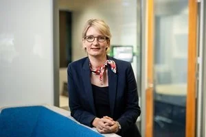 A woman with blonde hair, glasses, wearing a dark blazer and necklace, standing in an office or conference room with a blue table or counter.