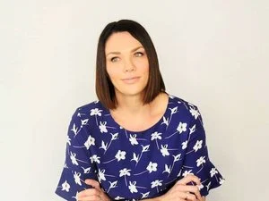 Portrait of a woman with dark hair in a blue dress with white floral patterns, standing against a plain background.