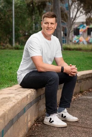 A young man with short hair wearing a white t-shirt, black jeans, and white sneakers sitting on a low stone wall in a park with trees and playground equipment in the background.