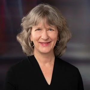 A woman with shoulder-length gray hair, wearing earrings and a black top, smiling at the camera.