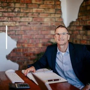 Man in glasses and suit sitting at a desk in front of a brick wall with a torn section.