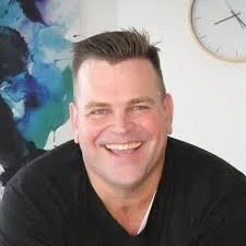 A smiling man with short dark hair, wearing a black shirt, sitting indoors with a colorful abstract painting and a clock on the wall behind him.