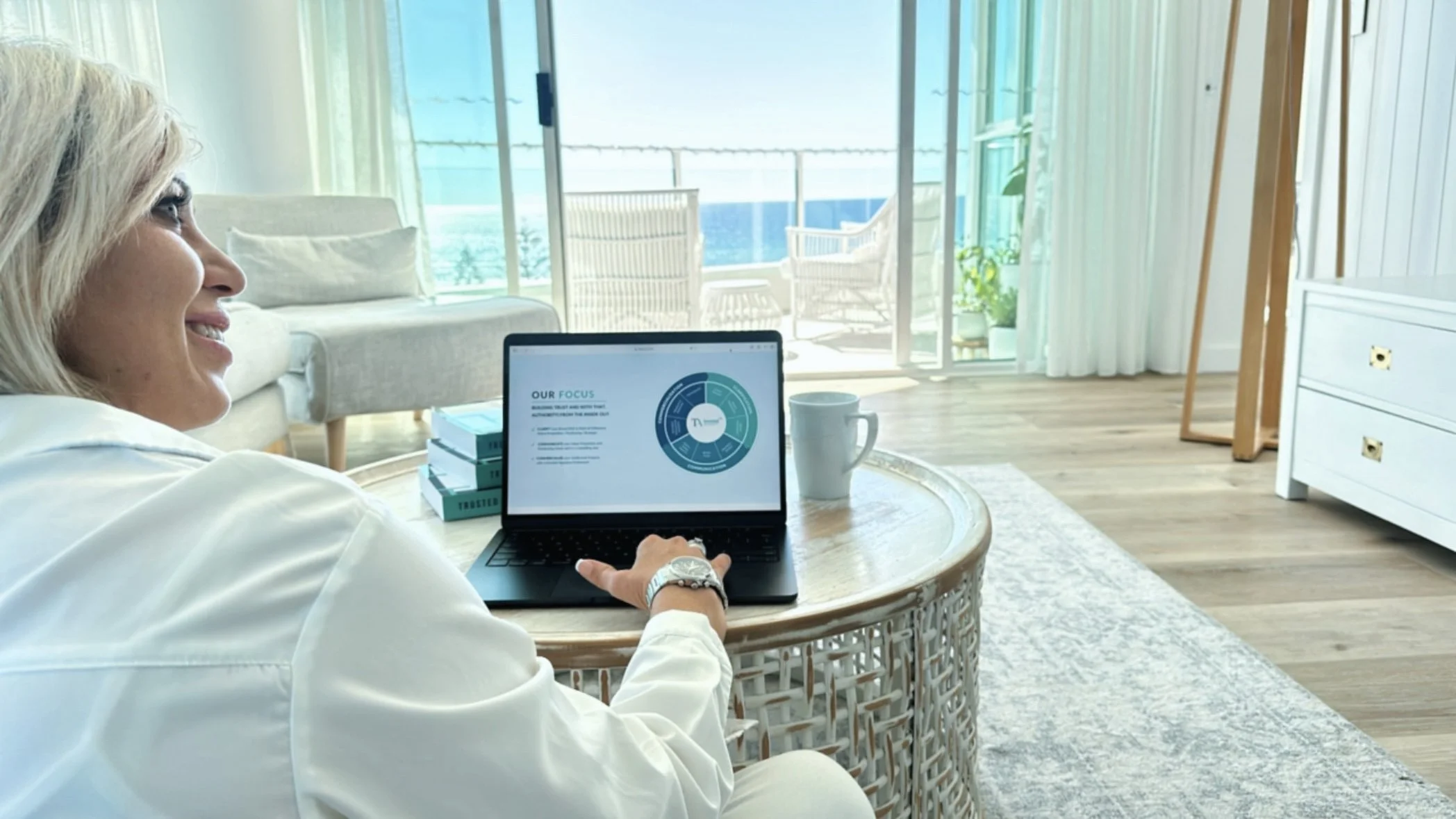 Woman sitting on a wicker coffee table, smiling, working on a laptop in a bright, modern living room with a view of the ocean outside