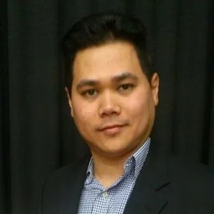 A man with dark hair in a suit and collared shirt, standing in front of a dark curtain background.