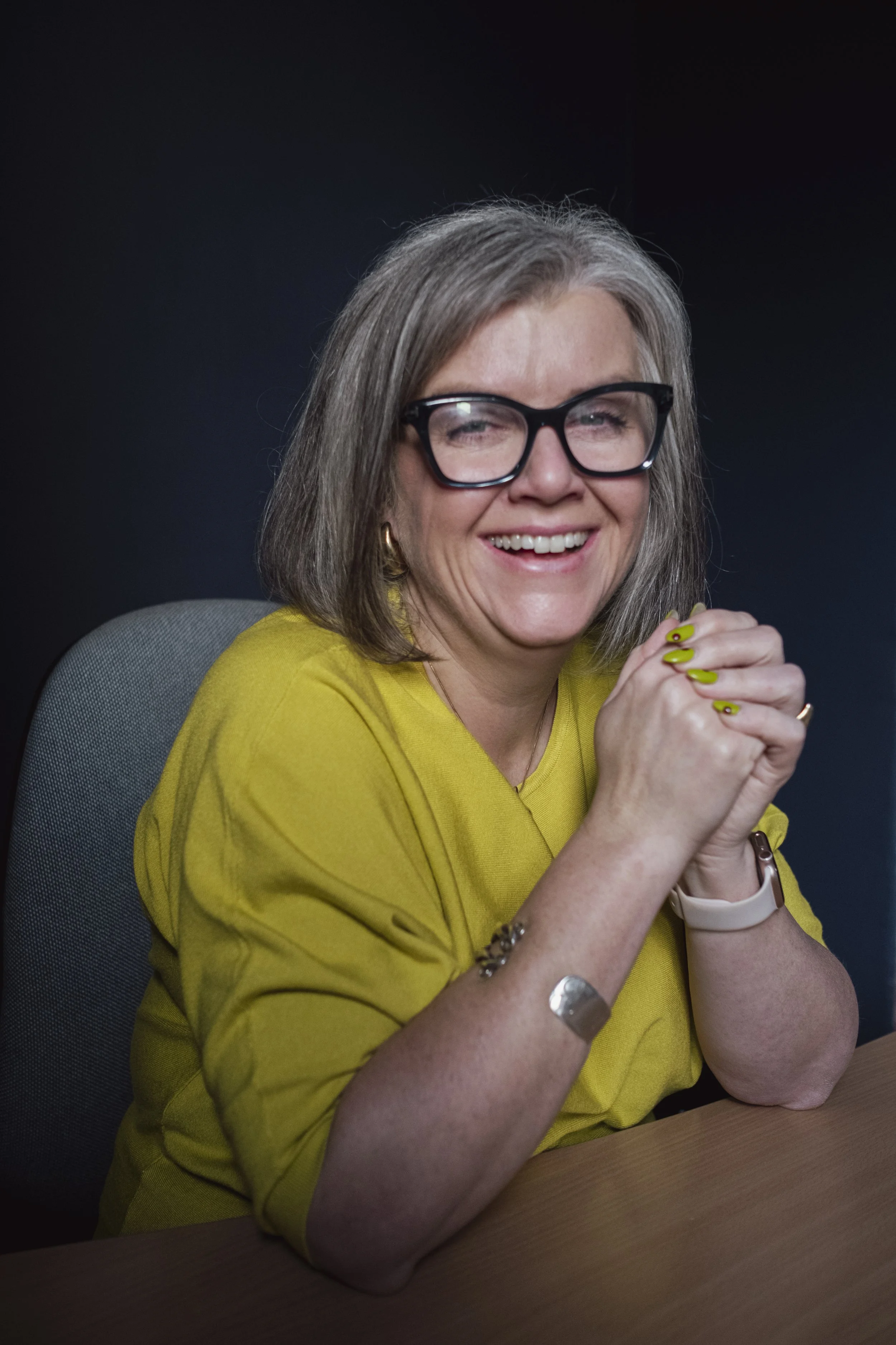 A smiling middle-aged woman with gray hair and black glasses, wearing a yellow top, sits at a wooden table with her hands clasped. She has yellow and silver nail polish, gold hoop earrings, a watch, and a bracelet, with a dark blurred background.