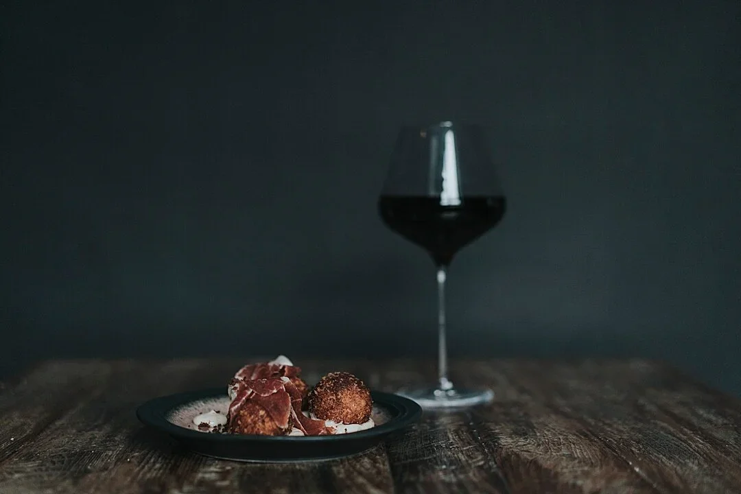 A glass of red wine with a dark background, placed on a rustic wooden surface, alongside a black plate with chocolate truffles and some edible decorations.