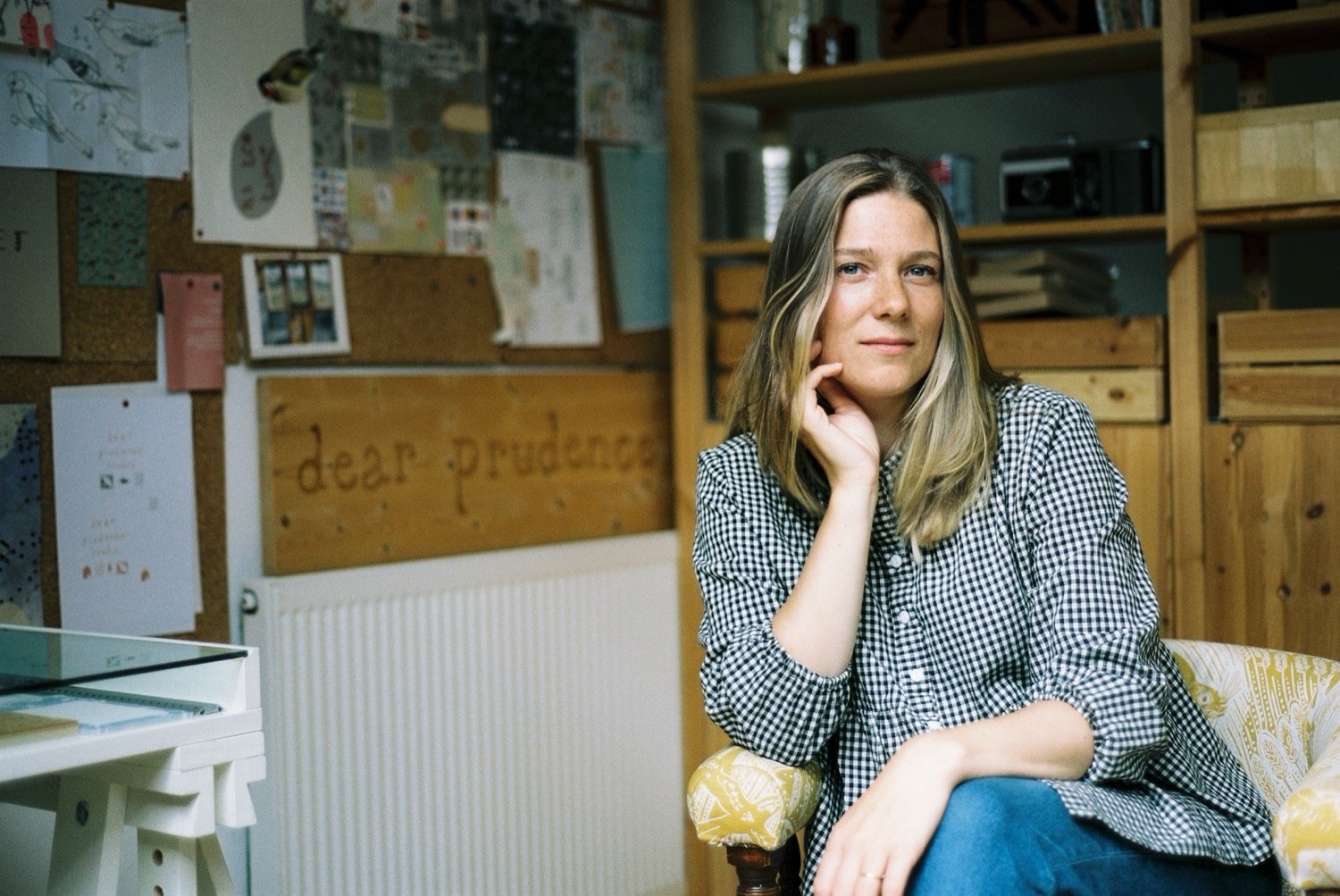 A woman with long blonde hair sitting in a patterned armchair in a room with wooden shelves and bulletin boards, wearing a checkered shirt and blue jeans.