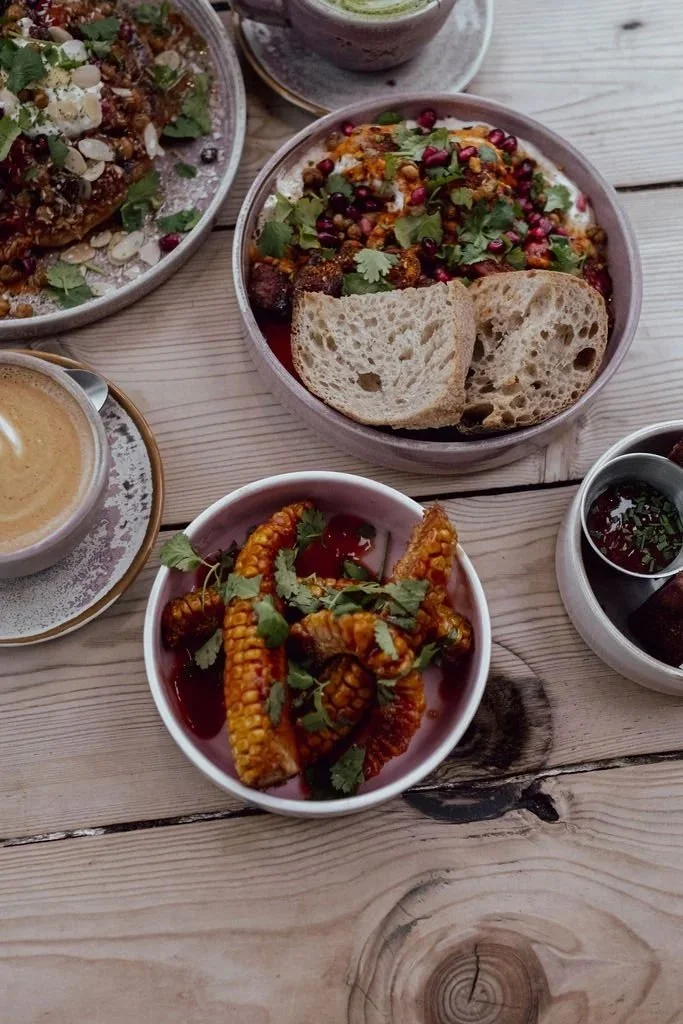A variety of Indian dishes including a bowl of curry with bread, a bowl of spiced vegetables garnished with herbs and pomegranate seeds, a bowl of fried vegetables in sauce, and small bowls of sauces or chutneys, all on a wooden table.