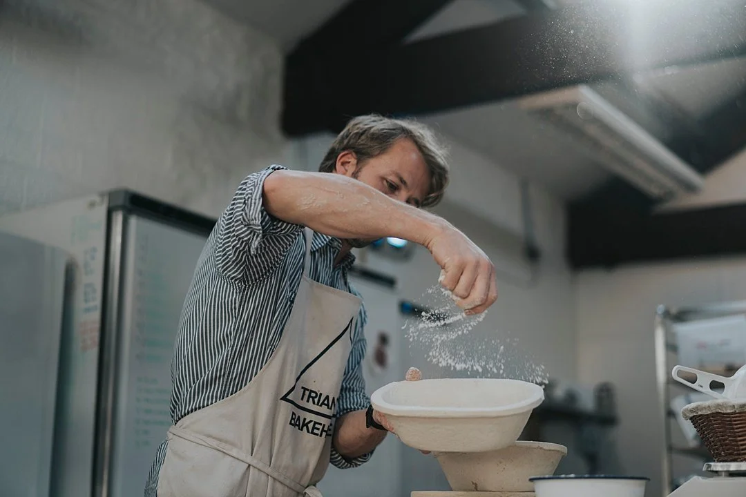 A man in a striped shirt and apron sprinkles flour into a bowl in a kitchen.